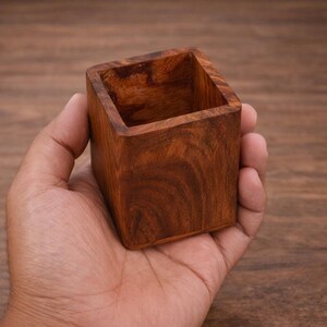 Puede incluir: Un peque&ntilde;o recipiente cuadrado de madera, de color marr&oacute;n oscuro con vetas visibles. El recipiente se sostiene en una mano, destacando su tama&ntilde;o compacto y est&eacute;tica natural. Ideal para objetos peque&ntilde;os o como pieza decorativa.