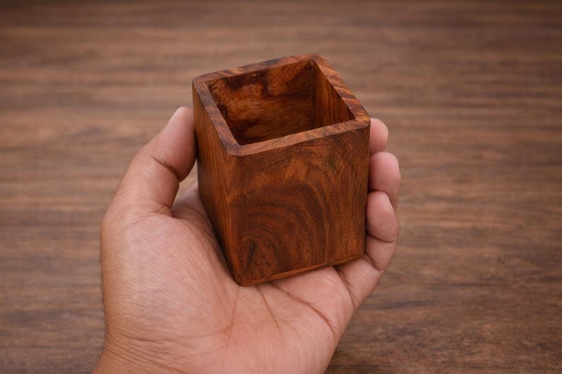 Puede incluir: Un peque&ntilde;o recipiente cuadrado de madera, de color marr&oacute;n oscuro con vetas visibles. El recipiente se sostiene en una mano, destacando su tama&ntilde;o compacto y est&eacute;tica natural. Ideal para objetos peque&ntilde;os o como pieza decorativa.