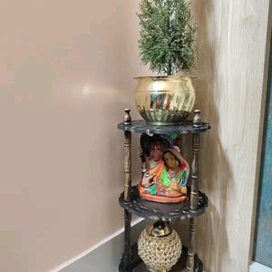 Pu&ograve; includere: Una mensola angolare in legno scuro a tre livelli con intagli decorativi. Il ripiano superiore contiene un vaso color oro con una pianta verde. Il ripiano centrale mostra una statuina colorata e il ripiano inferiore ha una sfera decorativa color oro.