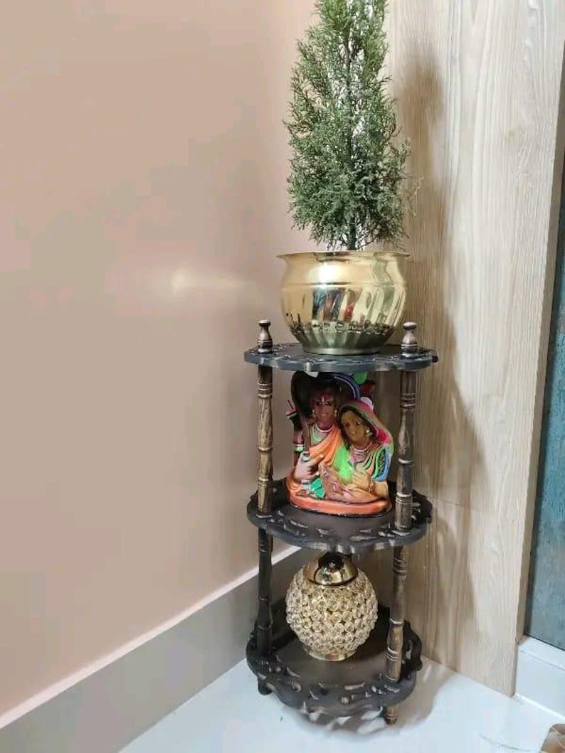 Pu&ograve; includere: Una mensola angolare in legno scuro a tre livelli con intagli decorativi. Il ripiano superiore contiene un vaso color oro con una pianta verde. Il ripiano centrale mostra una statuina colorata e il ripiano inferiore ha una sfera decorativa color oro.