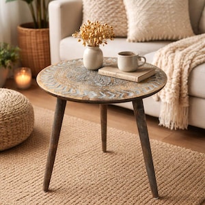 Puede incluir: Mesa auxiliar redonda de madera con diseño tallado y tres patas. Sobre la mesa, un jarrón con flores secas, una taza y un libro. Descansa sobre una alfombra de yute tejida, con un sofá beige y cojines al fondo.