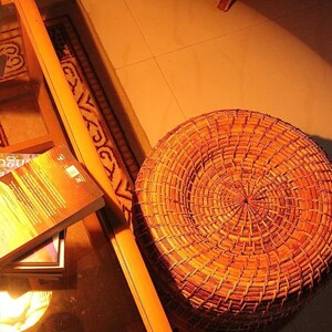 Peut inclure: Un tabouret rond en osier tress&eacute; aux tons bruns chauds. Le tabouret est pos&eacute; sur un sol carrel&eacute;, avec une table en verre et des livres en arri&egrave;re-plan. L'&eacute;clairage cr&eacute;e une atmosph&egrave;re chaleureuse et accueillante.