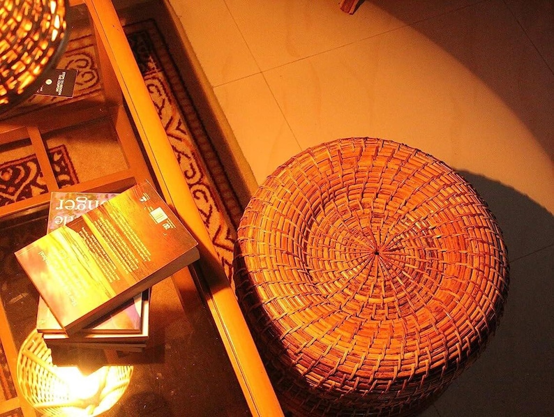 Peut inclure: Un tabouret rond en osier tress&eacute; aux tons bruns chauds. Le tabouret est pos&eacute; sur un sol carrel&eacute;, avec une table en verre et des livres en arri&egrave;re-plan. L'&eacute;clairage cr&eacute;e une atmosph&egrave;re chaleureuse et accueillante.