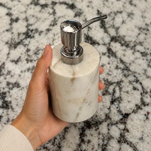 May include: A cylindrical soap dispenser crafted from white marble with natural grey veining. The dispenser features a polished chrome pump mechanism at the top, providing a functional and sleek design for dispensing liquid soap or lotion.