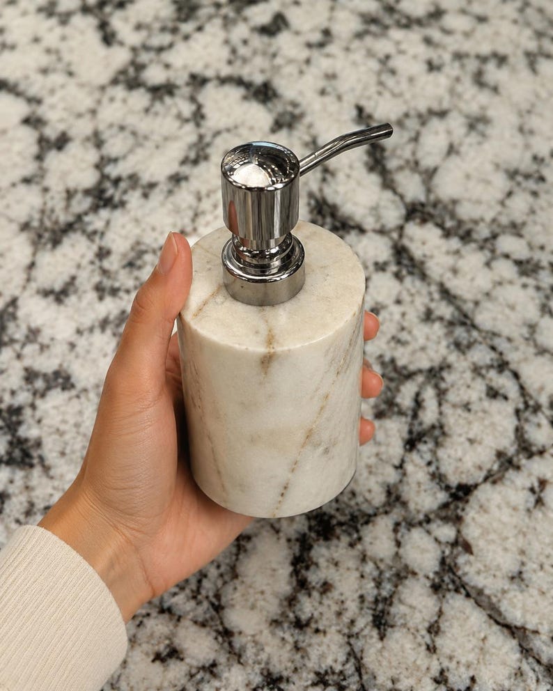 May include: A cylindrical soap dispenser crafted from white marble with natural grey veining. The dispenser features a polished chrome pump mechanism at the top, providing a functional and sleek design for dispensing liquid soap or lotion.