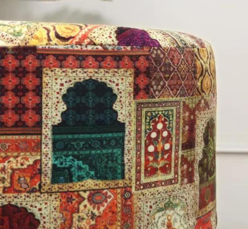 May include: Close-up of a round ottoman with a patchwork design. The fabric features a variety of patterns, including floral, geometric, and traditional rug motifs in shades of red, green, gold, and purple. The ottoman has a soft, cushioned appearance.
