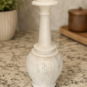 May include: A white marble vase with a smooth, polished finish. The vase features a rounded, bulbous base that transitions into a slender, elongated neck, topped with a wide, flat circular rim. The stone displays subtle grey veining throughout.