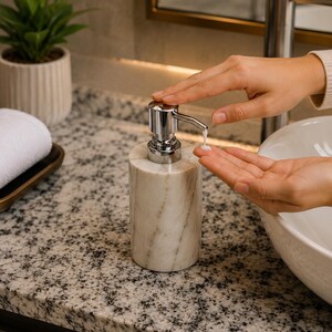 May include: A cylindrical soap dispenser crafted from white marble with grey veining, featuring a polished chrome pump mechanism at the top. The dispenser is designed for liquid soap and provides a smooth, functional aesthetic for a bathroom vanity.