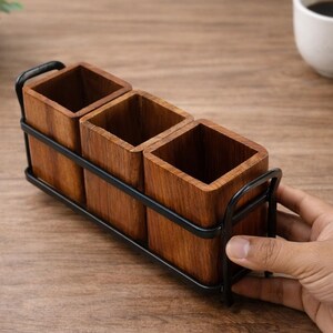 Puede incluir: Un portautensilios de madera con tres compartimentos cuadrados. El soporte est&aacute; hecho de madera de color marr&oacute;n claro y est&aacute; encerrado en un marco de metal negro. El soporte est&aacute; dise&ntilde;ado para colocarse sobre una mesa o encimera.