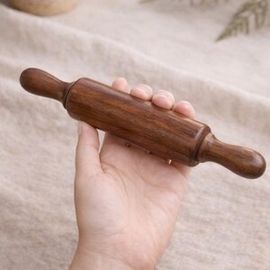 Puede incluir: Un rodillo de madera peque&ntilde;o con un acabado marr&oacute;n oscuro. El rodillo se sostiene en una mano, mostrando sus mangos lisos y redondeados y su cuerpo cil&iacute;ndrico. Ideal para hornear y hacer pasteles.