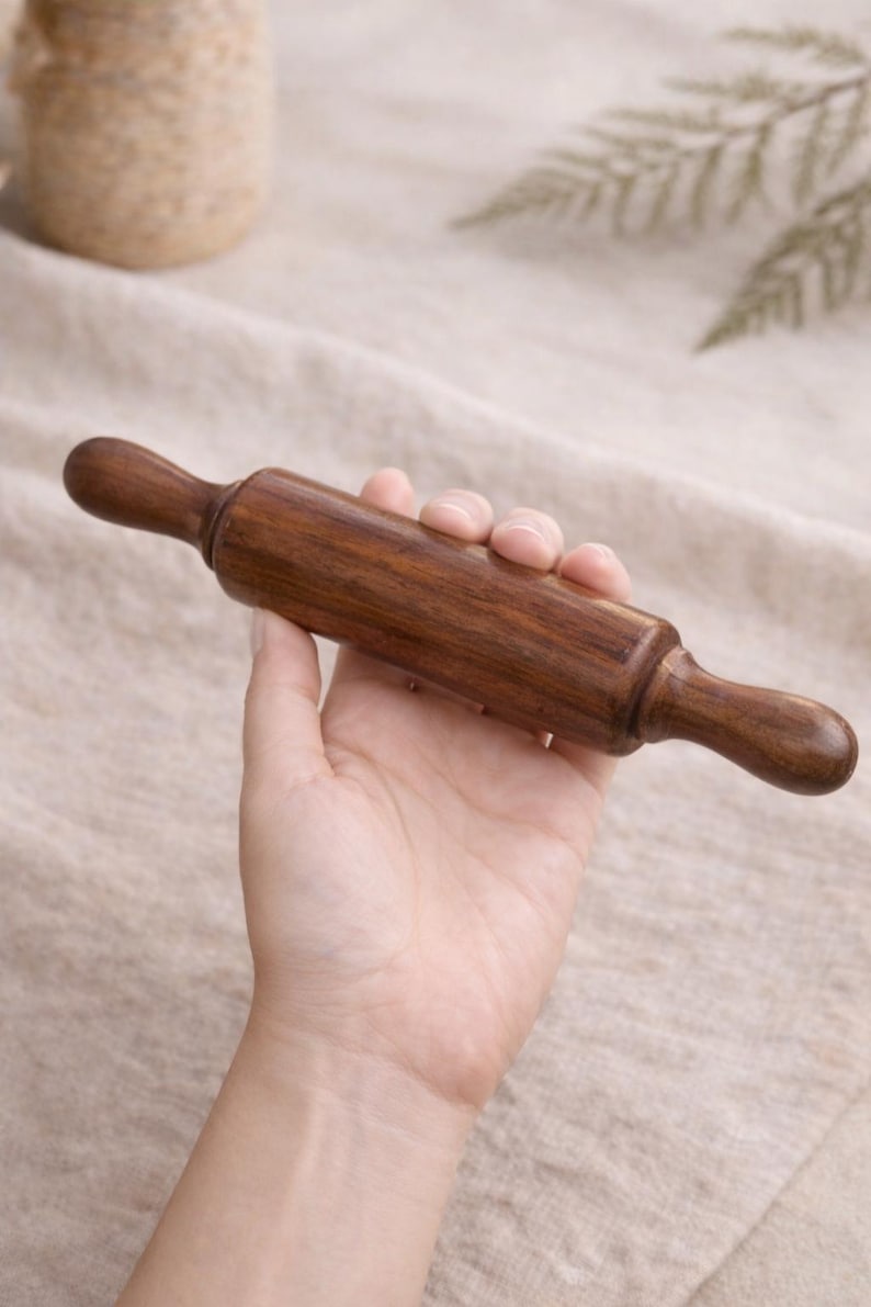 Puede incluir: Un rodillo de madera peque&ntilde;o con un acabado marr&oacute;n oscuro. El rodillo se sostiene en una mano, mostrando sus mangos lisos y redondeados y su cuerpo cil&iacute;ndrico. Ideal para hornear y hacer pasteles.