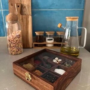 May include: A wooden spice box with a glass lid, filled with various spices, sits on a countertop. A glass oil dispenser, jars of spices, and a jar of cereal are also visible. Wooden cutting boards are in the background.