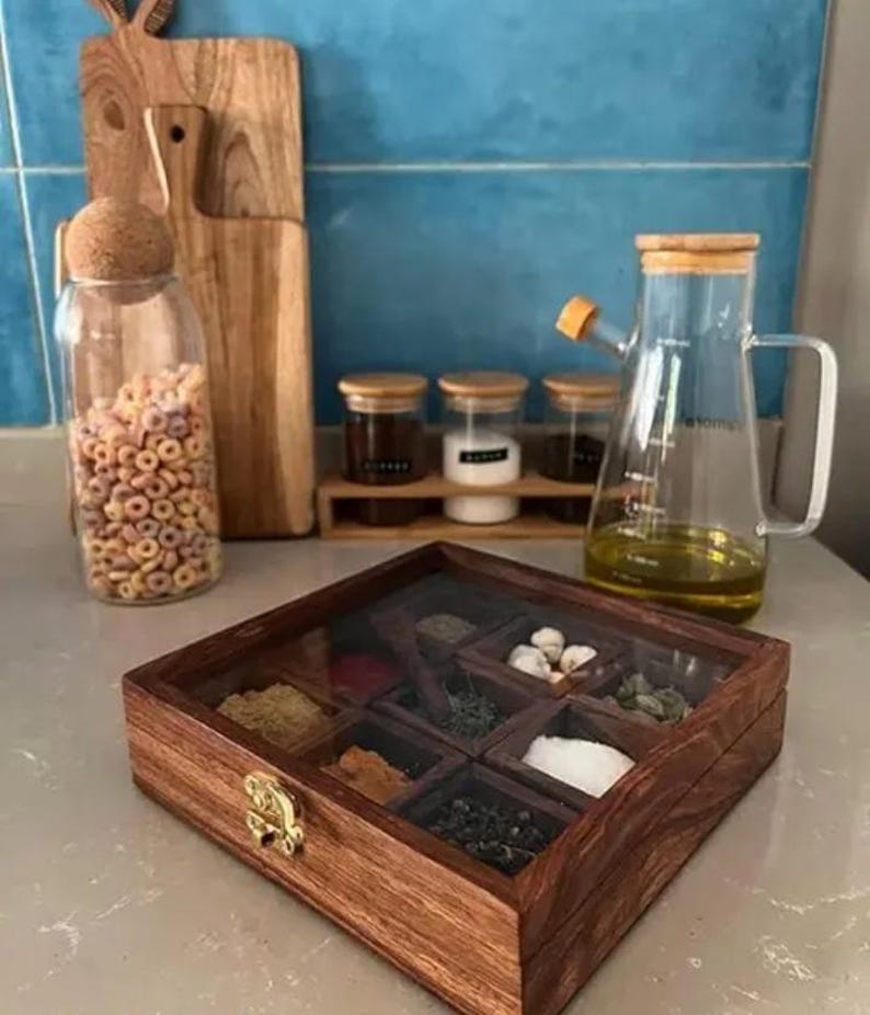 May include: A wooden spice box with a glass lid, filled with various spices, sits on a countertop. A glass oil dispenser, jars of spices, and a jar of cereal are also visible. Wooden cutting boards are in the background.