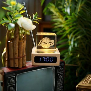 May include: A wooden digital clock with a clear cube featuring the Los Angeles Lakers logo. The clock displays the time 17:00 in blue digits. The cube is illuminated, and the clock sits on a vintage television.
