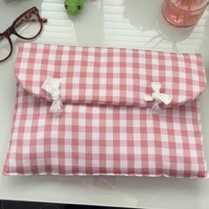 Puede incluir: Una funda para portátil con estampado de cuadros rosas y blancos y cierre de solapa. La funda se asegura con dos lazos de tela blanca. El diseño es de estilo campestre clásico.