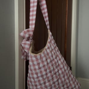 Può includere: Una borsa tote a fantasia gingham rosa e bianca con cinturino e fiocco abbinati. La borsa è appesa a una maniglia. La borsa ha una texture trapuntata e un interno morbido e di colore chiaro.