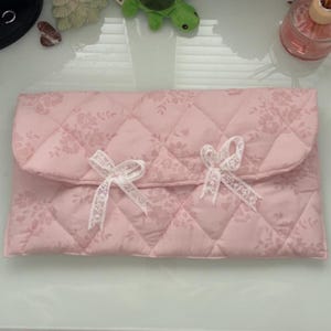 May include: A quilted pink clutch with a floral pattern and two white lace bows. The clutch has a flap closure and a diamond pattern. The overall theme is feminine and delicate.