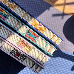 May include: Three hardback books stacked diagonally, featuring exposed Coptic stitch binding with decorative coloured thread patterns. The spines show cream-coloured pages and intricate, multi-coloured woven stitching in blue, pink, and orange hues.