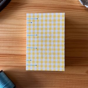 Può includere: Un piccolo taccuino cucito a mano con un motivo a quadretti giallo e bianco. Il taccuino è rilegato con filo azzurro. Una bobina di filo blu e altri oggetti artigianali sono sullo sfondo su una superficie di legno.