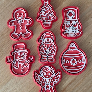 Peut inclure: Un ensemble de sept emporte-pièces en plastique rouge représentant des formes de Noël, dont un bonhomme en pain d'épices, un sapin, un casse-noisette, un bonhomme de neige, le Père Noël, une boule et un ange, avec des détails en relief blancs.
