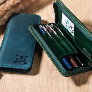 Puede incluir: Un estuche de cuero color turquesa con cierre de cremallera y un diseño en relieve de flechas, un sombrero, un bigote y las letras "K" y "S". El estuche está junto a un estuche verde abierto con bolígrafos dentro.