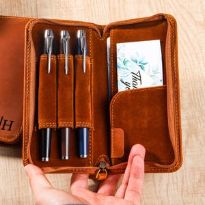 Puede incluir: Estuche de cuero marrón abierto con tres bolígrafos y una tarjeta que dice "Gracias". El estuche tiene cremallera y un bolsillo para la tarjeta. Se ve un cuaderno de cuero a juego con iniciales.