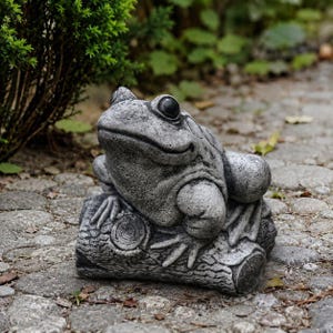 May include: A grey stone garden statue of a frog resting on a textured log. The frog has large, dark, prominent eyes and detailed skin texture, while the log features visible wood grain patterns and circular ends, creating a realistic appearance.