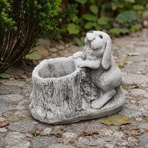 May include: A grey stone-effect planter shaped like a hollow tree stump with a sculpted rabbit leaning against the side. The rabbit has long ears and a textured surface, while the stump features realistic bark detailing and a circular opening.