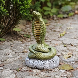 Puede incluir: Una figura de serpiente cobra verde con un patrón de escamas texturizado y vientre color canela, enroscada en posición erguida. La serpiente descansa sobre una base circular blanca con textura de veta de madera, creando un adorno de jardín detallado.