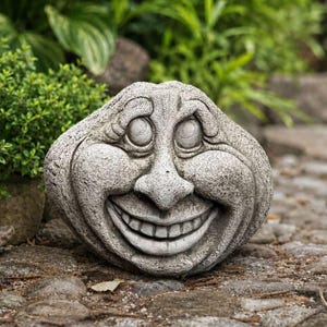 May include: A grey stone garden ornament shaped like a rounded face with a wide, toothy grin, prominent cheeks, and large, circular eyes. The surface has a textured, weathered appearance with deep grooves defining the facial features.