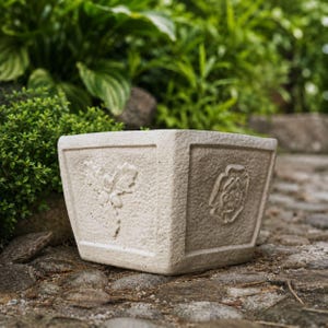 May include: A square, off-white stone planter with a textured, rough surface. The front face features a raised relief design of a cluster of holly leaves and berries, while the side face displays a raised relief of a stylised floral rose emblem.