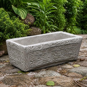 May include: A rectangular concrete planter with a textured surface that mimics the appearance of weathered wood grain. The planter features vertical panels on the corners and a horizontal wood-like pattern across the sides, finished in a light gray color.