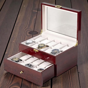 May include: A two-tiered, cherry-red wooden watch box with a glass top and a pull-out drawer. The interior features white padded compartments holding several wristwatches. The box is open, revealing the watches inside, and sits on a dark wood surface.