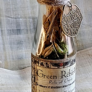May include: A clear glass bottle with a bronze-colored cap and a decorative metal tag. The bottle contains dried plants and moss. The label reads "Green Rebirth, Relic of Osiris, Jade Dragon Emporium."