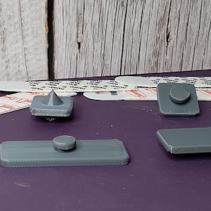 May include: A collection of gray plastic adhesive picture hanging hardware. The set includes various shapes, including rectangular and square pieces with circular and conical protrusions, and adhesive strips.