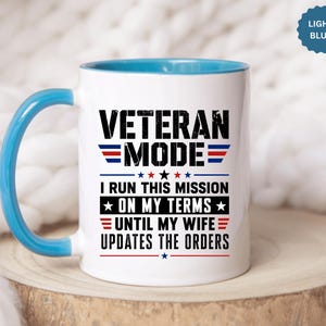May include: A white ceramic mug with a light blue handle and interior rim. The front features black text that reads VETERAN MODE I RUN THIS MISSION ON MY TERMS UNTIL MY WIFE UPDATES THE ORDERS, accented with red, white, and blue stars and stripes.