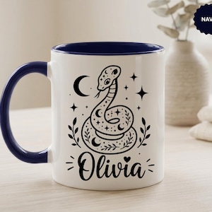 May include: A white ceramic mug with a navy blue interior and handle. The front features a black graphic illustration of a coiled snake surrounded by stars, a crescent moon, and leafy sprigs. The name Olivia is printed in black script below.