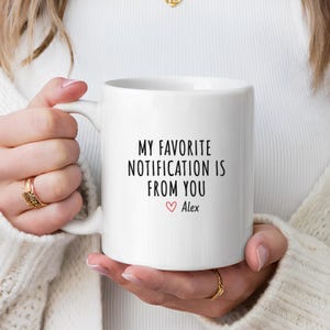Può includere: Tazza in ceramica bianca con la scritta "MY FAVORITE NOTIFICATION IS FROM YOU" in nero, con un cuore rosso e il nome "Alex" sotto. La tazza ha un manico curvo.