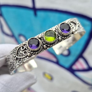 May include: A silver cuff bracelet featuring a textured, oxidized metal surface with intricate scrollwork patterns. The bracelet is set with three round, faceted gemstones: a deep purple stone on each end and a vibrant lime green stone in the center.