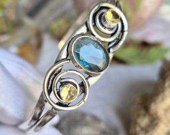 May include: A silver cuff bracelet featuring a central oval-cut blue gemstone flanked by two smaller round-cut yellow gemstones. The metal band is crafted with swirling, open-work metal loops that frame each of the three faceted stones securely.