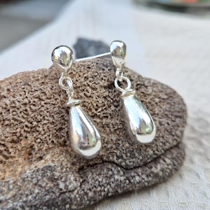 May include: A pair of polished silver drop earrings featuring a smooth, rounded stud top connected by a small jump ring to a larger, elongated teardrop-shaped pendant. The metal surface is highly reflective, showing a bright, metallic finish.