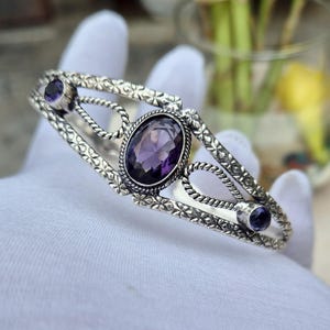 Può includere: Un bracciale a fascia in argento con una pietra ametista viola centrale a taglio ovale, affiancata da due pietre viola rotonde più piccole. La fascia in metallo presenta un intricato motivo floreale e dettagli a corda intrecciata.