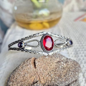 May include: A silver cuff bracelet featuring a central oval-cut red gemstone surrounded by a twisted metal border. The band has a textured floral pattern and includes two smaller dark purple circular stones set on either side of the centre.