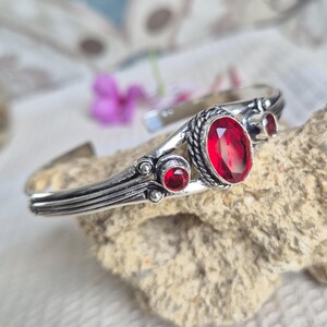 May include: A silver cuff bracelet featuring a large oval faceted red gemstone in the centre, flanked by two smaller round faceted red gemstones. The metal band has a textured, oxidised finish with decorative rope-like detailing around the stones.