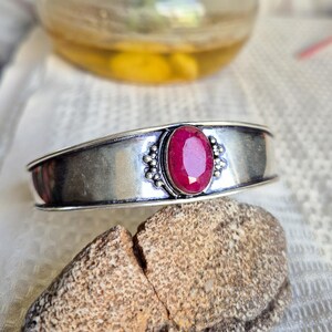 Könnte beinhalten: Ein silbernes Armband mit einem zentralen, oval facettierten, rosaroten Edelstein. Der Stein ist in eine dekorative Metallfassung mit kleinen perlenartigen Akzenten auf beiden Seiten eingefasst, die in das glatte, polierte Silberband übergeht.