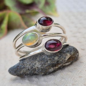 May include: A stack of three silver rings, each featuring a single oval-shaped gemstone. The top and bottom rings are set with deep red garnet stones, while the middle ring contains a translucent, iridescent opal with subtle green and yellow hues.