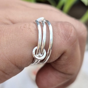 Könnte beinhalten: Ein silberfarbener Ring mit einem Mehrfach-Design, bestehend aus drei einzelnen, glatten und polierten Metallringen, die miteinander verbunden sind. Die Ringe sind abgerundet und haben eine reflektierende, metallische Oberfläche, die einen geschichteten Look erzeugt.