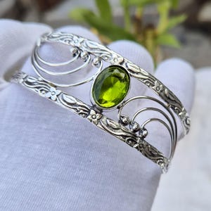 May include: A silver cuff bracelet featuring an oval-cut, lime green peridot gemstone at the centre. The metal band is decorated with embossed floral patterns and delicate, curved wire accents that extend from the central stone setting.