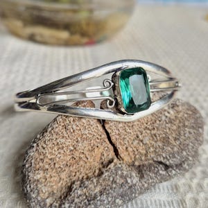 May include: A silver cuff bracelet featuring a central rectangular emerald-green gemstone. The metal band has a polished finish with decorative wire scrollwork details on either side of the stone, creating an open, airy design structure.