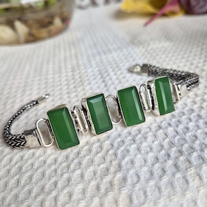 May include: A silver bracelet featuring four rectangular green stones set in metal frames. The bracelet has a woven chain design connecting the stone settings, creating a continuous link structure that secures with a metal clasp mechanism.